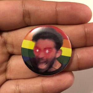 May include: A round button with a rainbow pride flag design. The center of the button features a close-up image of a person's face with red eyes and an open mouth.