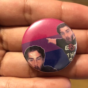 May include: A round button with a pink, purple, and blue background. The button features two men with dark hair and beards. The man on the right is pointing to the left and wearing a black shirt with the text "Eat the Rich" and an anarchy symbol. The man on the left is wearing a black shirt and glasses.