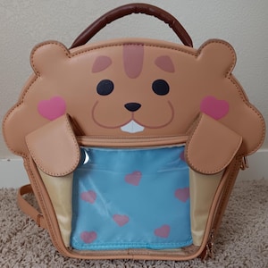 May include: A brown faux leather backpack shaped like a hamster with a clear plastic front pocket. The pocket has a blue lining with pink hearts printed on it. The hamster has a pink nose and pink cheeks.