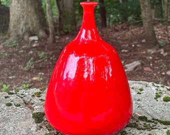 May include: A bright red ceramic vase with a rounded, bulbous shape and a narrow neck. The vase is sitting on a mossy rock.