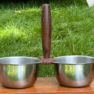 May include: A stainless steel double bowl set with a wooden handle. The bowls are round and have a smooth, shiny finish. The handle is dark brown and has a natural wood grain.