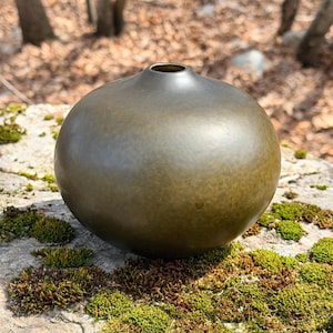 May include: A bronze-colored, spherical vase with a small opening at the top. The vase has a smooth, rounded shape and sits on a moss-covered rock. The background features blurred trees and fallen leaves, suggesting an outdoor setting.