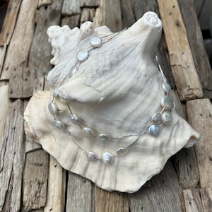 May include: A silver chain necklace with white round pearl-like beads. The necklace is layered and has a delicate design. The necklace is displayed on a white seashell.