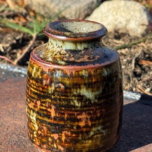 May include: A small, brown and green ceramic vase with a textured surface. The vase has a wide mouth and a narrow base.