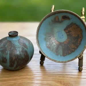 May include: A small, round, teal-colored ceramic vase with a narrow neck sits next to a teal and brown ceramic dish on a brass stand. The vase and dish have a mottled, textured appearance, suggesting a handcrafted quality. The objects are displayed on a wooden surface.