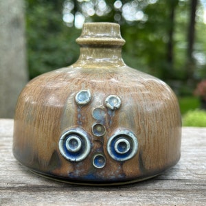 May include: A small, brown ceramic vase with a textured glaze and blue and white accents. The vase has a round body and a narrow neck.