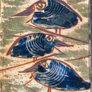 May include: A rectangular ceramic tile featuring a textured, mottled light green and beige background. Three stylized birds are depicted in a vertical row, each rendered in deep blue with dark brown outlines and intricate, swirling line details.