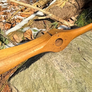 May include: A wooden airplane propeller with red tips. The propeller is made of light brown wood and has a hole in the center. It is lying on a rock with a brown and green background.