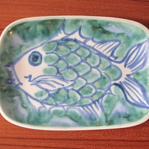 May include: A ceramic dish with a blue and green fish design. The fish has a white body and blue fins and scales. The dish is a light green color with a white rim.