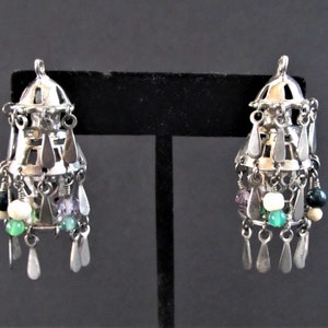 May include: Silver-toned chandelier earrings with a tiered design. Each earring features dangling teardrop-shaped charms and colorful beads in green, white, and purple. The earrings are displayed against a dark background.