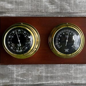 May include: A vintage barometer and hygrometer set. The instruments have black faces with white markings and gold-colored bezels, mounted on a dark wood plaque. The barometer is on the left, and the hygrometer is on the right.