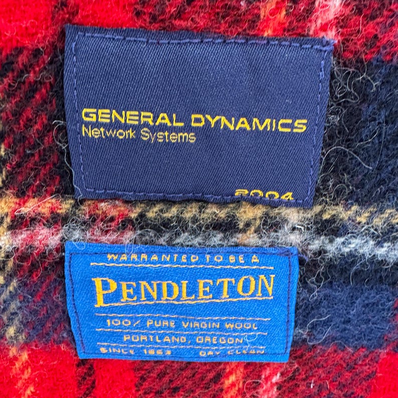 May include: Close-up of two blue fabric labels on a red, black, and yellow plaid background. The top label reads "GENERAL DYNAMICS Network Systems" in gold lettering. The bottom label says "PENDLETON" in large blue letters.