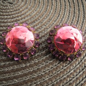 May include: Pair of vintage clip-on earrings. Each earring features a large, faceted pink center stone surrounded by smaller purple stones. The setting is gold-toned. The earrings are displayed on a textured, brown surface.