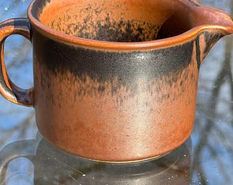 May include: A brown ceramic creamer with a black glaze and a speckled finish. The creamer has a handle and a spout.