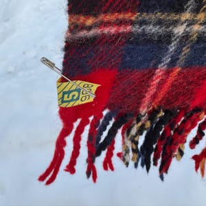 May include: A close-up shot of a red, black, and navy plaid scarf with fringed edges. A safety pin holds a yellow tag with green text. The scarf has a textured look, possibly wool or a similar fabric.