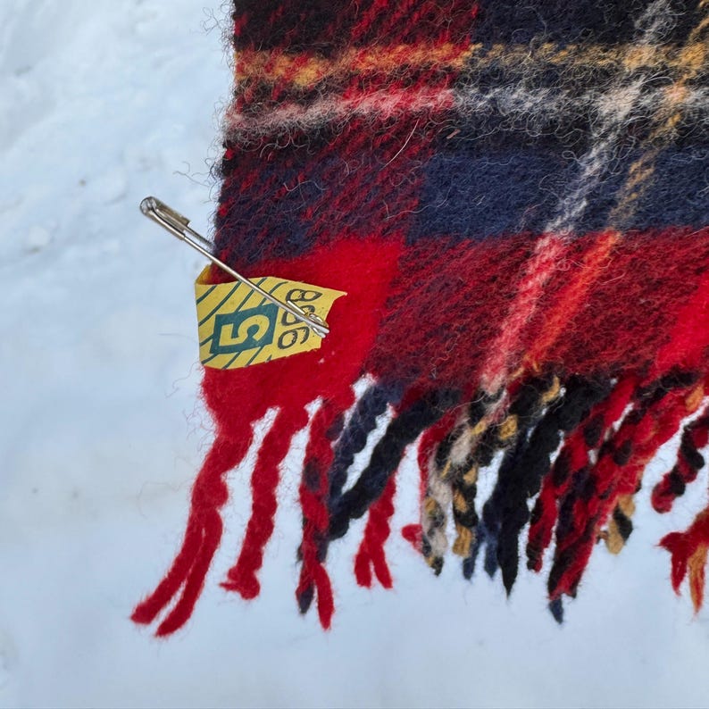 May include: A close-up shot of a red, black, and navy plaid scarf with fringed edges. A safety pin holds a yellow tag with green text. The scarf has a textured look, possibly wool or a similar fabric.