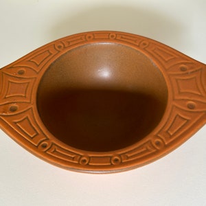 May include: A brown ceramic bowl with a geometric design around the rim. The bowl has a rounded shape and two small handles on either side.