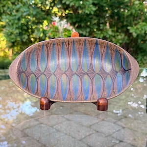May include: An oval ceramic dish featuring a repeating pattern of blue and green leaf-like shapes against a textured brown background. The dish is supported by a small, dark brown wooden stand with two rounded feet, elevating the piece.