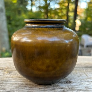 May include: A small, round, brown ceramic vase with a dark brown rim. The vase has a glossy finish and is sitting on a wooden surface.
