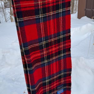 May include: A red, navy blue, and black plaid scarf with fringe. The scarf is hanging outside on a metal bar. The scarf has a blue tag with white text.