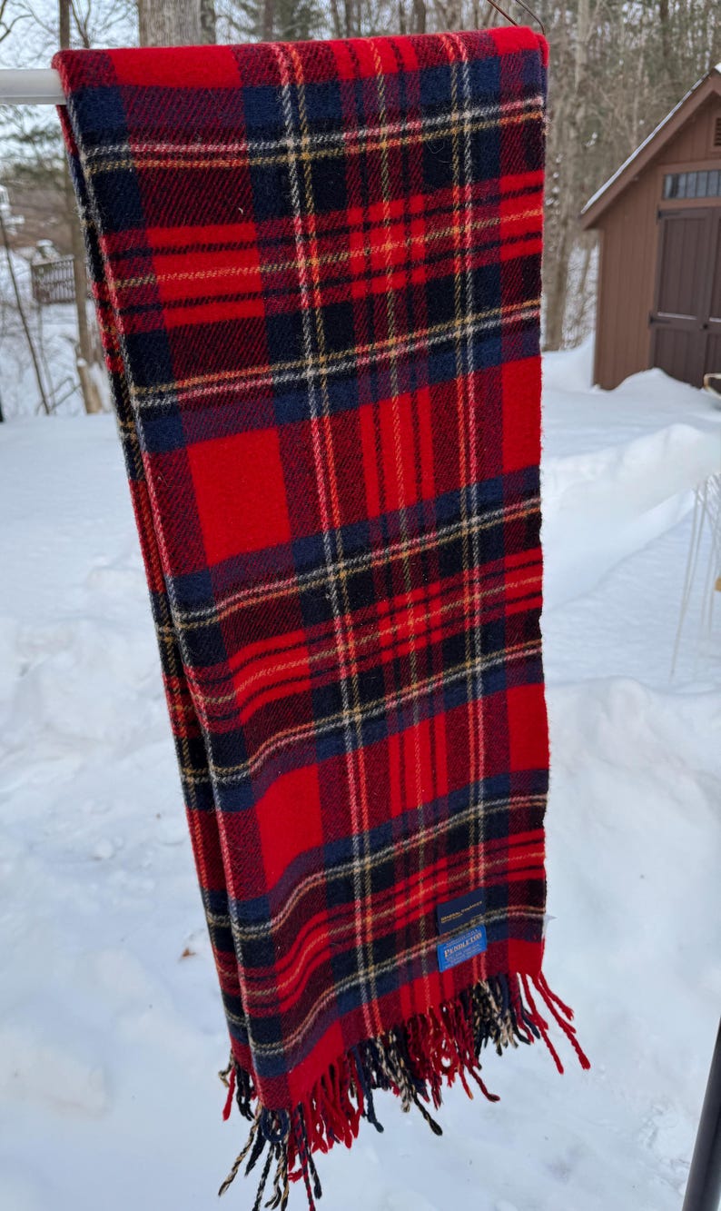 May include: A red, navy blue, and black plaid scarf with fringe. The scarf is hanging outside on a metal bar. The scarf has a blue tag with white text.