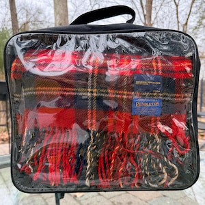 May include: A Pendleton wool blanket in a black carrying case. The blanket features a red, black, and gold plaid design with a fringed edge. The carrying case has a black handle and a clear plastic front. The blanket has two blue labels with text.