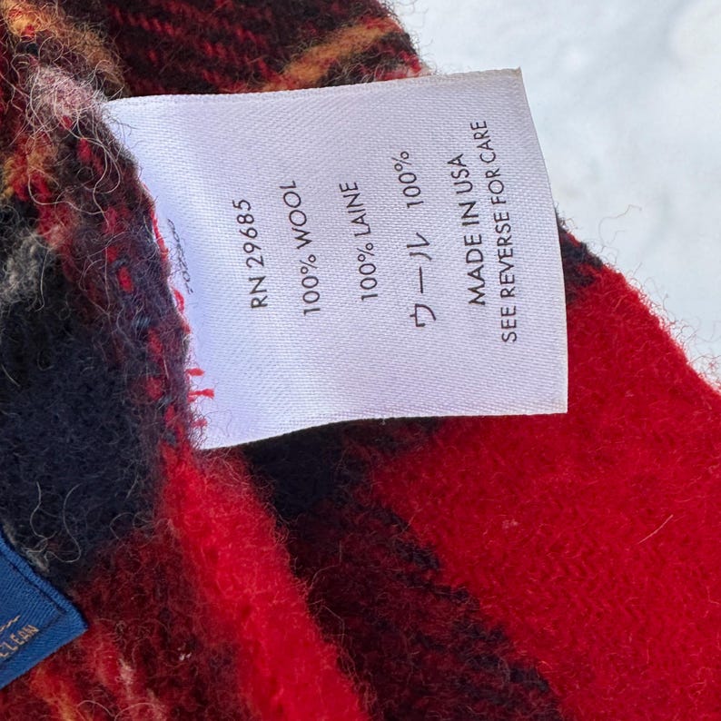 May include: A close-up shot of a label on a red, black, and yellow plaid fabric. The label displays text including "100% WOOL", "100% LAINE", and "MADE IN USA SEE REVERSE FOR CARE". The fabric is likely wool.
