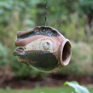 May include: A ceramic birdhouse shaped like a fish with brown, white, and black glaze. The birdhouse has a hole in the front for birds to enter.