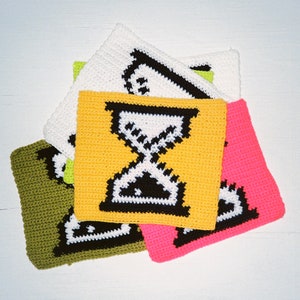 May include: Four crocheted squares with different colored backgrounds. The squares are stacked on top of each other. The top square is white with a black hourglass design. The second square is yellow with a black hourglass design. The third square is green with a black hourglass design. The bottom square is pink with a black hourglass design.