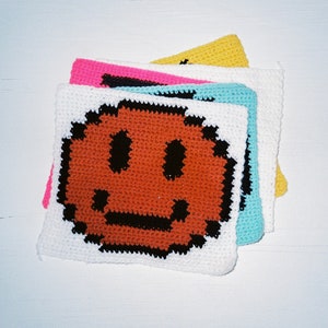 Puede incluir: Una pila de cuatro cuadrados de crochet, cada uno con un fondo de color diferente. El cuadrado superior es blanco con una cara sonriente marrón tejida en el centro. Los otros cuadrados son rosa, azul y amarillo.