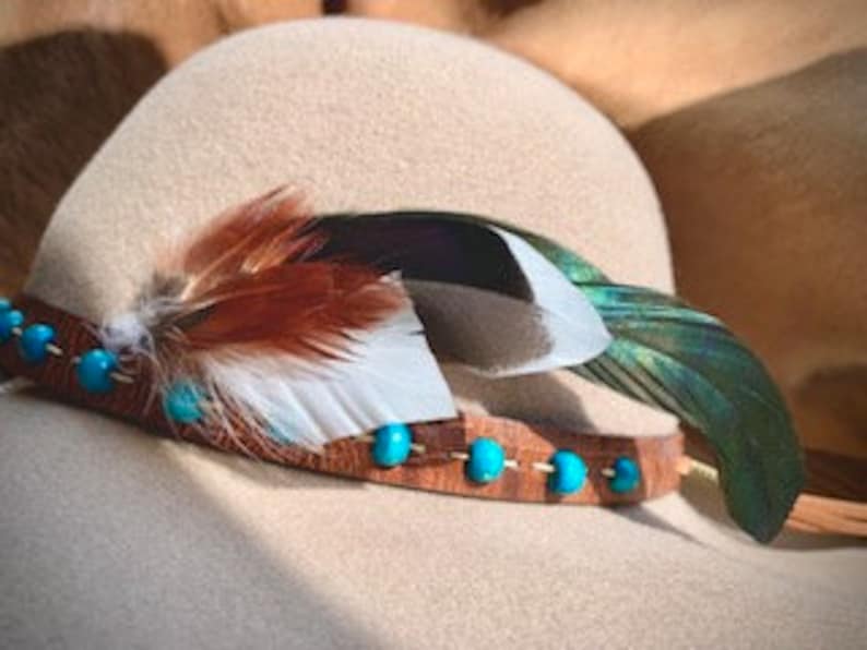 Puede incluir: Una banda de sombrero de cuero marr&oacute;n con cuentas turquesa y una pluma marr&oacute;n, blanca y verde.