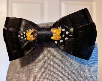 May include: A black bow tie with a gold and white feather accent.