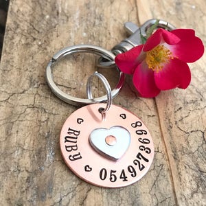 Może przedstawiać: Identyfikator dla zwierząt domowych z miedzi i aluminium z zawieszką w kształcie serca. Identyfikator jest grawerowany imieniem "RUBY" i numerem telefonu "0549273698".