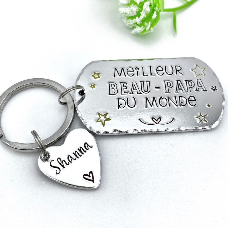 May include: A silver-toned metal keychain featuring a rectangular tag engraved with the text MEILLEUR BEAU-PAPA DU MONDE and small star accents. A smaller heart-shaped charm is attached to the ring, engraved with the name Shanna and a heart.