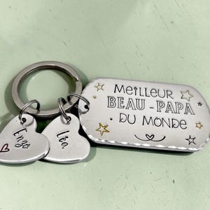 May include: A silver-toned metal keychain featuring a rectangular tag engraved with the text MEILLEUR BEAU-PAPA DU MONDE, surrounded by small star icons. Two smaller heart-shaped charms are attached, each engraved with the names Enzo and L&eacute;a.