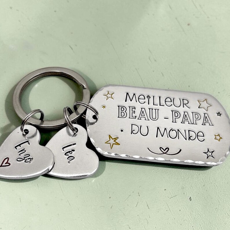 May include: A silver-toned metal keychain featuring a rectangular tag engraved with the text MEILLEUR BEAU-PAPA DU MONDE, surrounded by small star icons. Two smaller heart-shaped charms are attached, each engraved with the names Enzo and L&eacute;a.