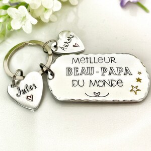May include: A silver-toned metal keychain featuring a rectangular tag engraved with the text MEILLEUR BEAU-PAPA DU MONDE and three small stars. Two heart-shaped charms are attached to the ring, one engraved with Jules and one with Aurore.