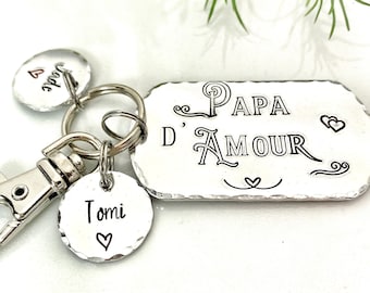 May include: A silver keychain featuring a rectangular tag with the words "Papa D'Amour" in black script. It includes round tags with the names "Tomi" and "Jade", each accompanied by a heart symbol. The keychain is equipped with a clasp for attachment.