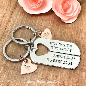May include: Two silver heart-shaped keyrings with a split design. Each keyring has a small heart charm with the initials "M&S" engraved on it. The keyrings are engraved with coordinates. The keyrings are on a wooden surface with pink flowers.