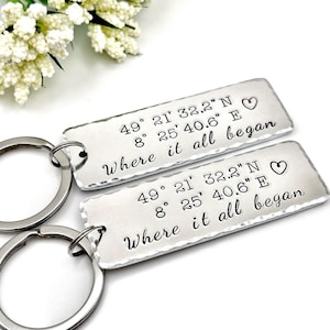 Puede incluir: Dos llaveros rectangulares de metal plateado con bordes martillados. Cada llavero está grabado con las coordenadas 49 21 32.2 N 8 25 40.6 E, un pequeño símbolo de corazón y el texto Where it all began en una fuente cursiva negra.