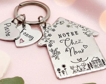 May include: A silver-toned metal keychain featuring a house-shaped charm engraved with the text "NOTRE Chez Nous" alongside floral, cat, dog, and house illustrations. Two smaller heart-shaped charms are attached, engraved with the names "Pierre" and "Fanny" with a small red heart symbol.
