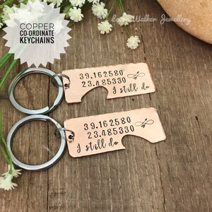 May include: Two copper keychains with matching coordinates and the text "I still do" engraved on each. The keychains are attached to silver key rings.
