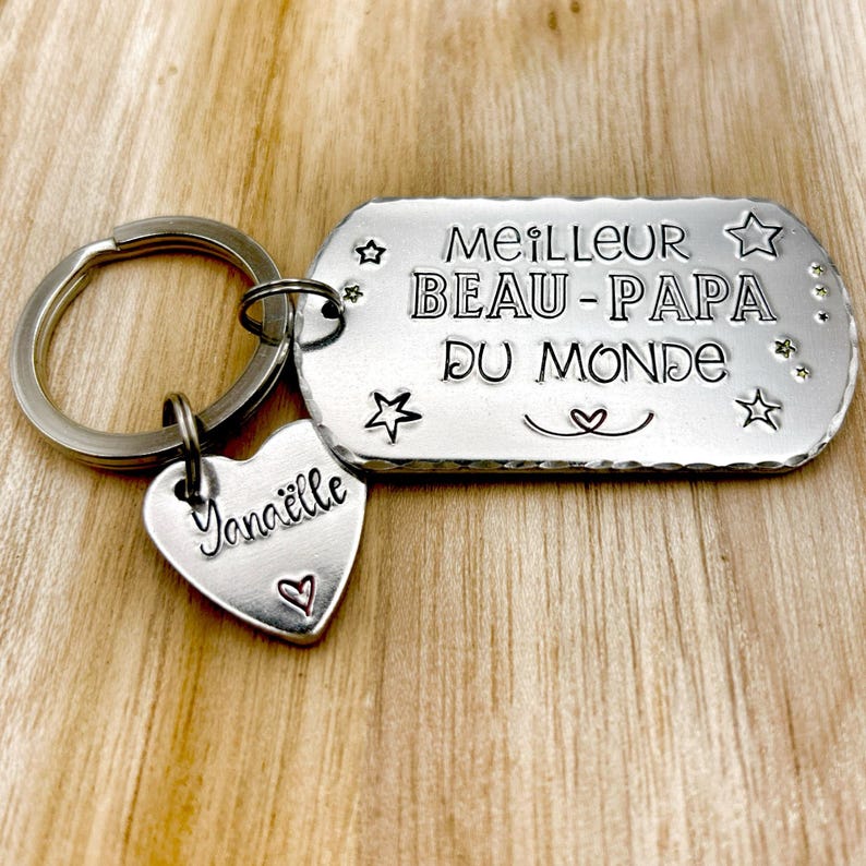 May include: A silver-toned metal keychain featuring a rectangular tag stamped with the text MEILLEUR BEAU-PAPA DU MONDE, star icons, and a heart symbol. A smaller heart-shaped charm is attached, engraved with the name Yana&euml;lle and a small heart.