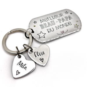 May include: A silver-toned metal keychain featuring a rectangular tag engraved with the text MEILLEUR BEAU-PAPA DU MONDE and star icons, accompanied by two heart-shaped charms engraved with the names Mila and Elise, each featuring a small heart symbol.
