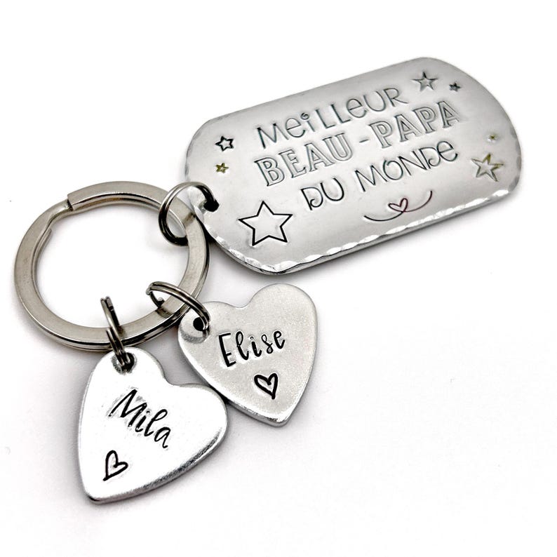 May include: A silver-toned metal keychain featuring a rectangular tag engraved with the text MEILLEUR BEAU-PAPA DU MONDE and star icons, accompanied by two heart-shaped charms engraved with the names Mila and Elise, each featuring a small heart symbol.
