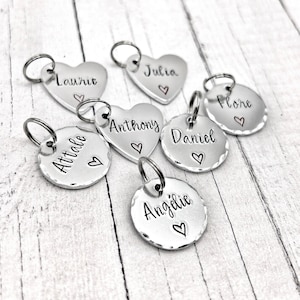 Puede incluir: Una colección de dijes de metal plateado en formas de corazón y circulares, cada uno con una argolla. Los dijes están estampados a mano con los nombres Laurie, Julia, Anthony, Daniel, Flore, Attale y Angélie, junto a un pequeño símbolo de corazón.