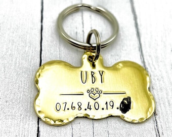 May include: A gold-toned metal pet ID tag shaped like a bone with a hammered edge. The tag is engraved with the name UBY, a small paw print heart icon, and the numerical sequence 07.68.40.19. A silver-colored metal split ring is attached to the top.