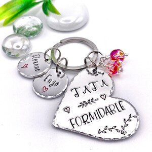 Może przedstawiać: Srebrny brelok w kształcie serca z wygrawerowanym napisem "TATA A FORMIDABLE". Brelok ma również dwa mniejsze srebrne kółka z wygrawerowanymi nazwami "Roxane" i "Enza". Brelok jest ozdobiony dwoma małymi różowymi i pomarańczowymi koralikami szklanymi.