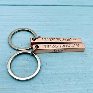 Op de afbeelding: Twee roségoudkleurige metalen staafsleutelhangers met zilverkleurige ringen. De bovenste staaf is gegraveerd met 13° 13' 37.524" E. De onderste staaf is gegraveerd met 52° 35' 24.824" N en de naam Talia gevolgd door een klein hartje.