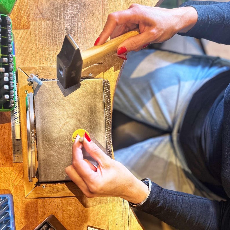 May include: A person using a hammer to shape a piece of metal on a workbench. The metal is on a gray surface with a yellow circle in the center. The person is wearing a black shirt and a bracelet.